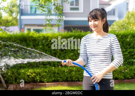 Asiatische Frau glücklich in Haus Hinterhof Sommergarten, mit Gartenschlauch Spritzwasser auf dem Rasen und Baum Blätter während des Aufenthalts zu Hause mit kostenlos Stockfoto