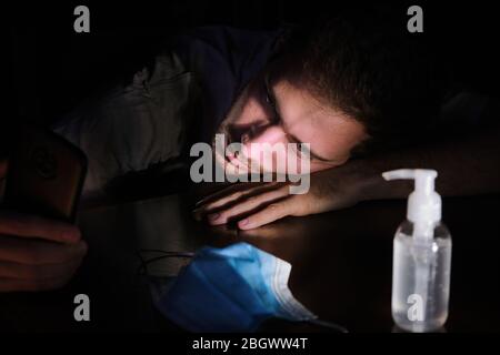 Ein Arzt erschöpft ruhend auf dem Tisch in der Nacht mit dem Handy-Bildschirm Beleuchtung, der Kinnriemen und der Gel-Alkohol während der covid 19 Pandemie Stockfoto