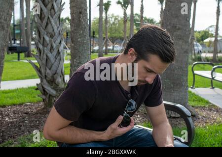 Ein Mann mit rotem Hemd und blauer Jeans entspannt sich auf einer Parkbank und spielt mit seiner Fliegerbrille und schaut auf einen hellen sonnigen Tag auf einer Parkbank herab Stockfoto