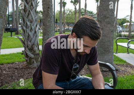 Ein Mann mit rotem Hemd und blauer Jeans entspannt sich auf einer Parkbank und spielt mit seiner Fliegerbrille und schaut auf einen hellen sonnigen Tag auf einer Parkbank herab Stockfoto