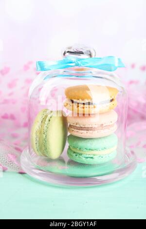 Sanfte bunte Makronen in Glas Glocke und Milchglas auf Farbe Holztisch, auf hellem Hintergrund Stockfoto