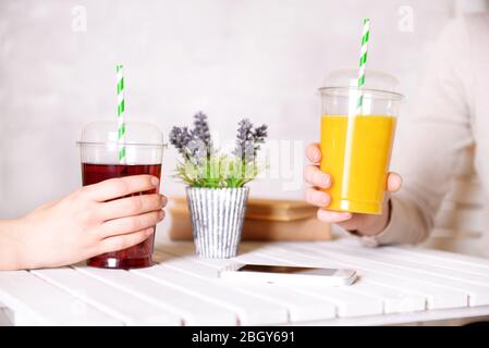 Zwei weibliche Hände mit Fast Food Tassen Saft auf Holztisch mit Pflanze auf hellen Wandhintergrund Stockfoto