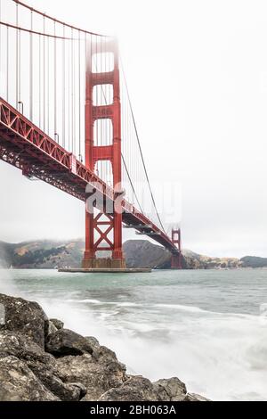 Die Golden Gate Brücke in einem nebligen Tag, wie von Fort Point aus gesehen, mit Felsen und brechenden Wellen im Vordergrund