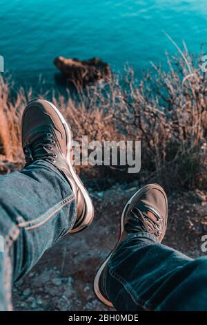 Beine des Menschen in Jeans und Schuhe hängen von Rand der Klippe über das Meer und Bäume Stockfoto