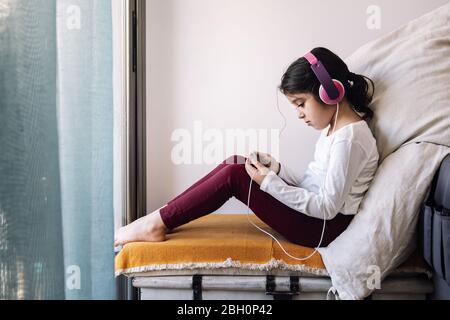 Kleines Mädchen sitzt vor ihrem Fenster mit Kopfhörern Videos auf dem Telefon, Home Entertainment für Kinder Konzept Stockfoto