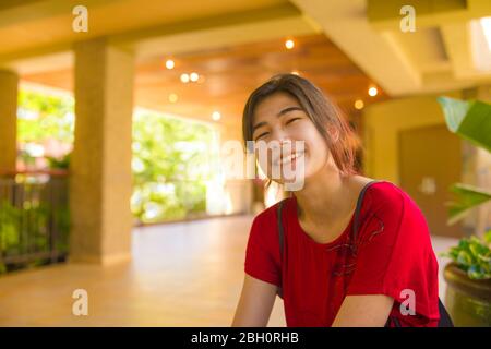 Lächelndes biracial asiatische kaukasische Teenager-Mädchen oder junge Frau sitzt in leeren Outdoor Open-Air-Lobby im tropischen sonnigen Hawaii, trägt rotes Hemd Stockfoto