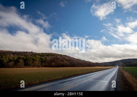 Eine asphaltierte, nasse Straße führt an einem Farmfeld vorbei in niedrige Hügel, unter einem blauen Himmel mit weißen Wolken im Westen von New York, USA Stockfoto