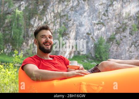 Lächelnder junger Mann Hipster mit einer Tasse Kaffee im Urlaub entspannt auf orangefarbenem Sofa. Freizeitkonzept, Camping, Sommerurlaub. Stockfoto