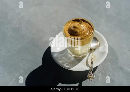 Dalgona Instant-Kaffee Schlagsahne Getränk mit Schokoladenpulver auf der Oberseite in Glas. Hartes Sonnenlicht, Betonhintergrund Stockfoto