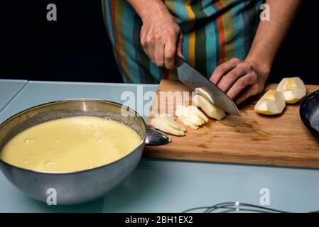 Arbeiten in der Küche.Detail der Hände schneiden Apfelstücke. Fruchtpüree. Stahlschale. Apfelkuchen kochen. Farbige Schürze. Küchentisch Dunkel schwarz Hintergrund Stockfoto