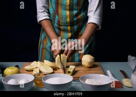 Arbeiten in der Küche. Halbbildfotografie. Detail der Hände schneiden Apfel in Stücke. Bunte Schürze. Schneidebrett. Kochmesser. Schwarzer Rücken Stockfoto