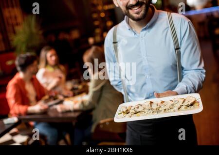 Lächelnder junger Kellner, der Teller mit Fleischgericht im Restaurant trägt Stockfoto