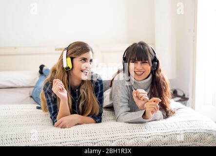 COVID-19-Sperre. Selbstbewusste und optimistische Frauen besten Freundinnen lachen online Musik hören singen und tanzen zusammen im Bett zu Hause in isol Stockfoto