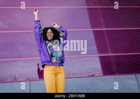 Junge Frau mit urbanem Look jubelt vor rosa Glaswand Stockfoto