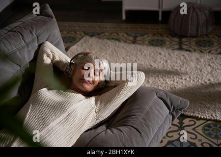 Porträt einer älteren Frau mit Kopfhörern, die sich zu Hause auf der Couch entspannen Stockfoto