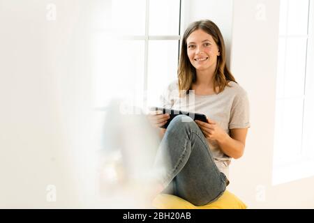 Lächelnde junge Brünette Frau zu Hause sitzen auf Fensterbank und hält ihr Tablet Stockfoto