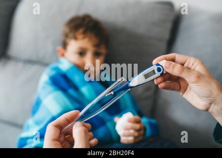 Mutter Hand nimmt Digitalthermometer, während sich Sohn auf Couch im Hintergrund wartet Stockfoto