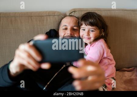 Glücklicher Vater und Tochter, die mit dem Handy auf der Couch sitzen, einen Videoanruf zur Familie machen Stockfoto