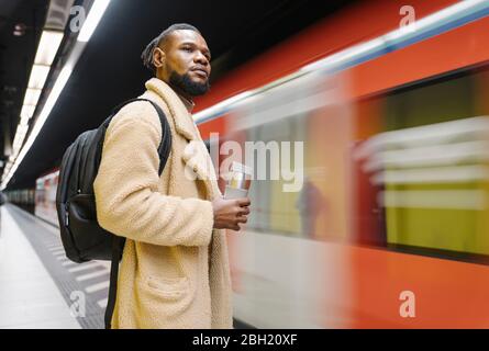 Stilvoller Mann mit wiederverwendbarem Becher und Kopfhörern in einer U-Bahn-Station Stockfoto