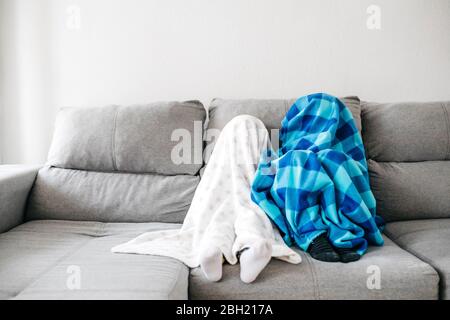 Das kleine Mädchen und ihr Bruder sitzen Seite an Seite auf der Couch zu Hause und verstecken sich unter Decken Stockfoto