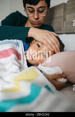 Porträt eines kranken Jungen, der im Bett liegt, während seine Mutter seine Stirn berührt Stockfoto