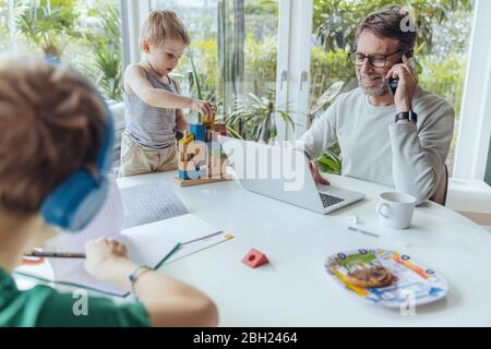 Vater spricht am Telefon, während Söhne auf dem Tisch spielen Stockfoto