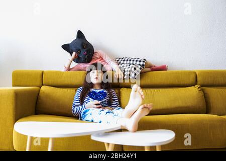 Zwei gelangweilte Mädchen sitzen auf der Couch, Fernsehen, eine trägt Katzenmaske Stockfoto