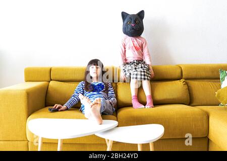 Zwei gelangweilte Mädchen sitzen auf der Couch, Fernsehen, eine trägt Katzenmaske Stockfoto