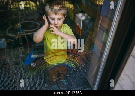 Porträt eines kleinen Jungen am Telefon hinter Balkontür stehend Stockfoto