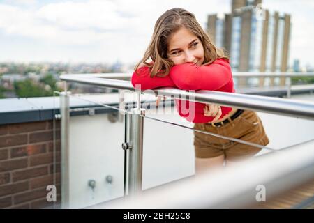 Porträt einer lächelnden jungen Frau, die auf dem Geländer auf dem Dach, London, Großbritannien, eaning Stockfoto