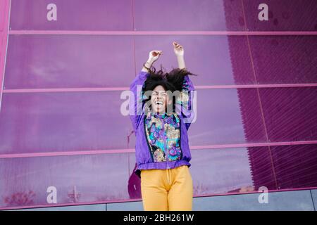 Junge Frau mit urbanem Look jubelt vor rosa Glaswand Stockfoto