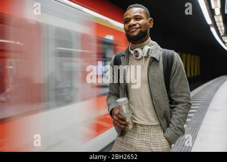 Stilvoller Mann mit wiederverwendbarem Becher und Kopfhörern in einer U-Bahn-Station Stockfoto