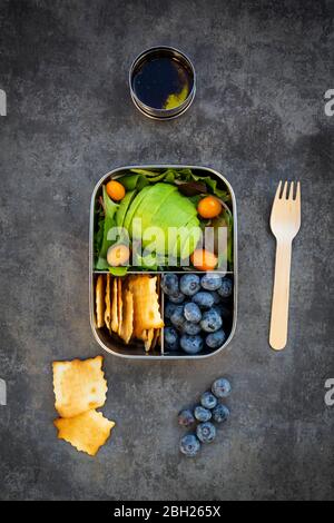 Schüssel Salatdressing und Lunchbox mit in Scheiben geschnittenen Avocado, gelben Tomaten, Cracker, Heidelbeeren und grünem Salat Stockfoto