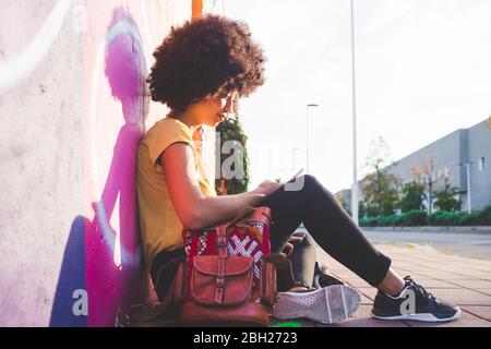 Junge Frau mit Afro-Frisur sitzt an der Graffiti-Wand und mit dem Smartphone Stockfoto
