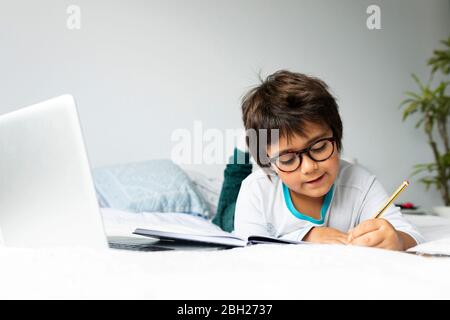 Portrait eines kleinen Jungen auf dem Bett liegend mit Laptop Hausaufgaben machen Stockfoto