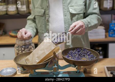 Nahaufnahme eines älteren Mannes hinter der Theke in seinem Laden, der getrocknete Lavendelblüten in einen Papierbeutel gießt Stockfoto