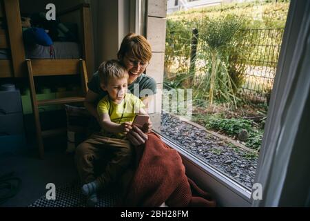 Mutter und kleiner Sohn sitzen zu Hause auf dem Boden mit dem Smartphone Stockfoto