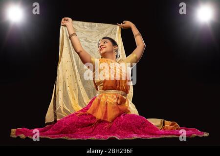 Kathak Tänzerin sitzt anmutig und tritt vor einem dunklen Hintergrund auf. Stockfoto