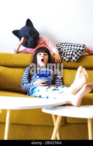 Zwei gelangweilte Mädchen sitzen auf der Couch, Fernsehen, eine trägt Katzenmaske Stockfoto