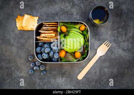 Schüssel Salatdressing und Lunchbox mit in Scheiben geschnittenen Avocado, gelben Tomaten, Cracker, Heidelbeeren und grünem Salat Stockfoto