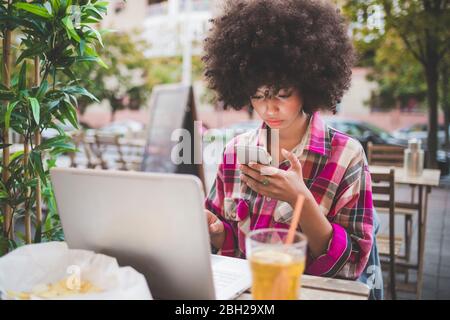 Junge Frau mit Afro-Frisur mit Smartphone und Laptop in einem Café im Freien in der Stadt Stockfoto