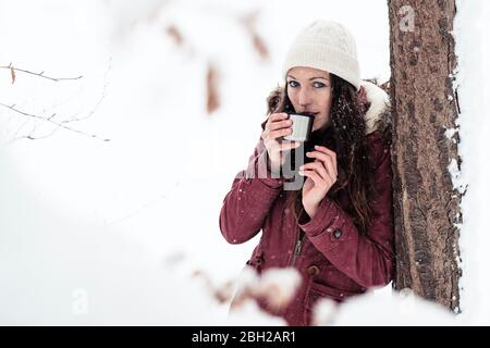 Porträt einer jungen Frau, die im Winter an Baumstamm lehnt und heißes Getränk trinkt Stockfoto