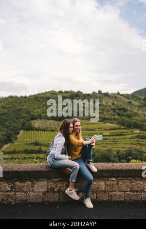 Zwei junge Frauen, die auf einer Mauer in ländlicher Landschaft sitzen und ein Selfie machen, Greve in Chianti, Toskana, Italien Stockfoto