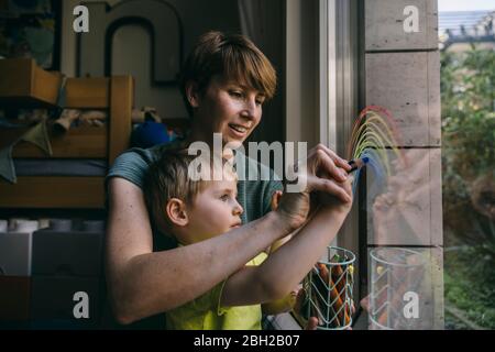 Mutter und kleiner Sohn zeichnen Regenbogen auf Fenster zu Hause Stockfoto