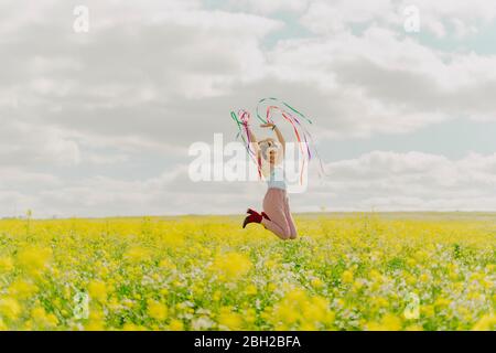 Glückliche junge Frau, die im Frühling mit bunten Bändern auf einer Blumenwiese springt Stockfoto