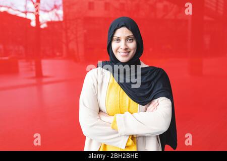 Porträt einer lächelnden jungen Frau, die Hijab vor einer roten Glasscheibe trägt Stockfoto
