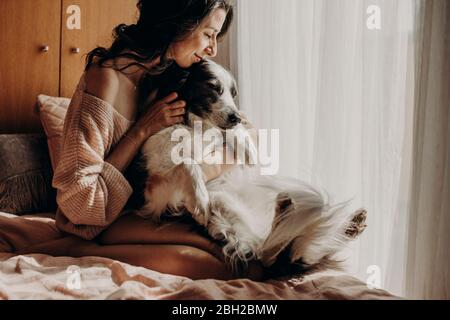 Glückliche Frau, die zu Hause auf dem Bett saß und ihren Hund streichelte Stockfoto