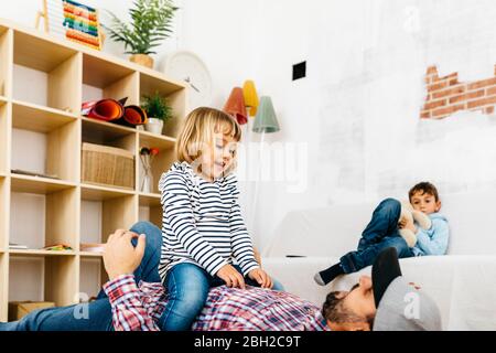 Tochter sitzt auf ihrem Vater, der auf dem Boden liegt und zu Hause spielt Stockfoto