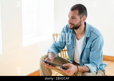Junger Mann zu Hause sitzt in einem Holzsessel vor dem Fenster mit Tablet Stockfoto