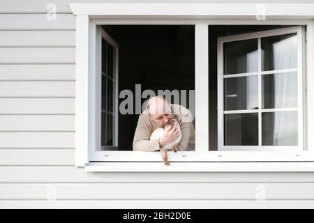 Reifer Mann mit Katze, die aus dem Fenster seines Hauses lehnt Stockfoto